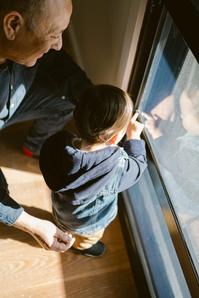 Dad watching over toddler showing him a bug he found just outside the sun-lit window where they sat