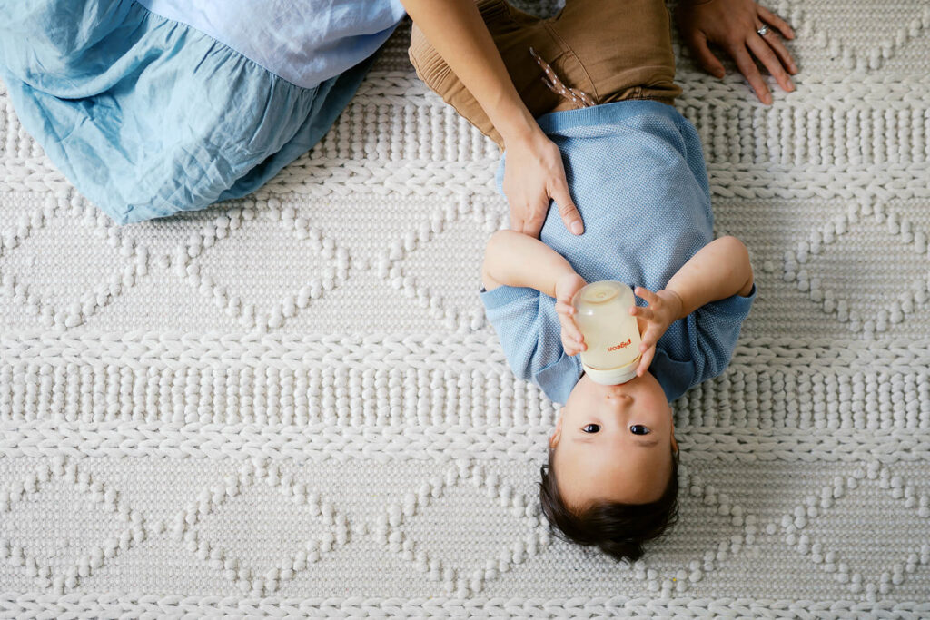 close up candid of toddler drinking milk from the bottle with mum's hand in the frame
