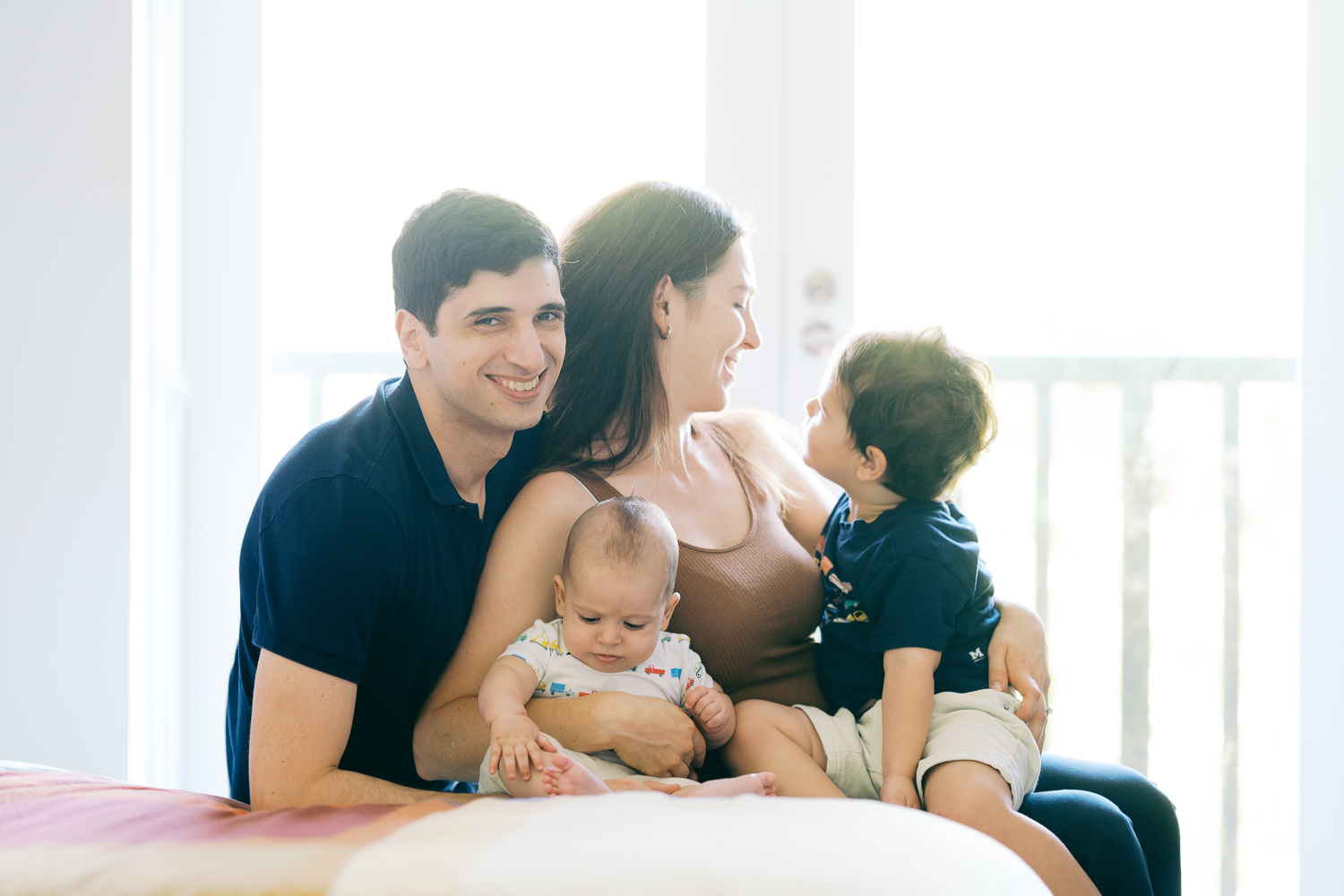 Family of 4 playing and cuddling with toddler and baby on the bed at their home