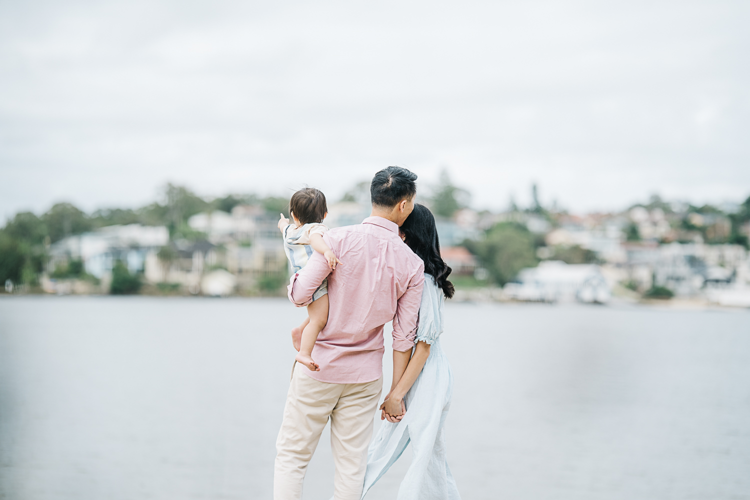 Young family enjoying each other near the water. Dad lovingly giving mum a kiss on the forehead while holding her hand and carrying their baby on the other hand